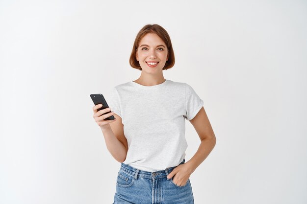 Retrato de mujer sonriente con smartphone, charlando en las redes sociales, de pie con el teléfono celular contra la pared blanca