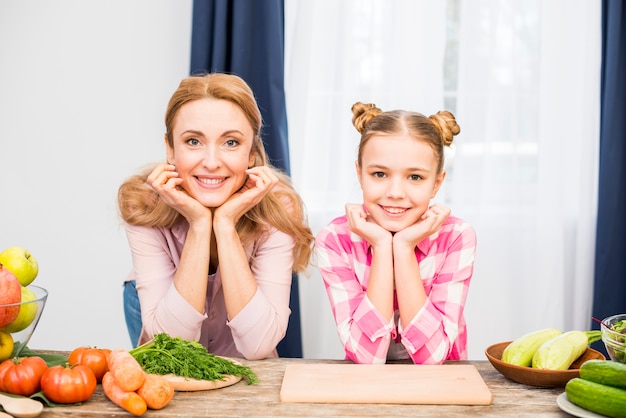 Retrato de una mujer sonriente y su madre que se inclinan sobre la mesa con la cabeza en la mano mirando a la cámara