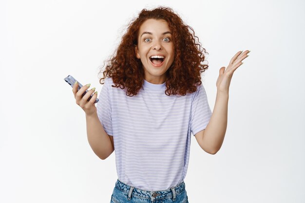 Retrato de mujer sorprendida y feliz con el pelo rojo, usando el teléfono móvil y celebrando, sonriendo sorprendida, reacción a una buena noticia impresionante, ganando el premio en el teléfono inteligente en blanco.