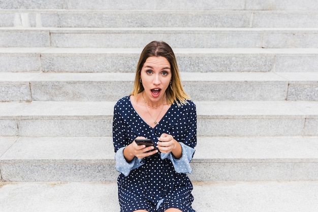 Foto gratuita retrato de una mujer sorprendida que se sienta en la escalera que sostiene el teléfono móvil