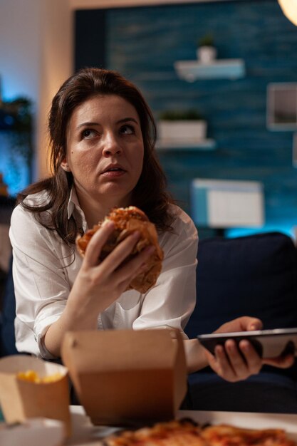 Retrato de mujer sosteniendo una sabrosa cena para llevar hamburguesa navegando en el teléfono inteligente con aspecto aburrido sentado en el sofá en la sala de estar de casa. Persona que tiene un menú de comida rápida a domicilio mientras navega por las redes sociales.