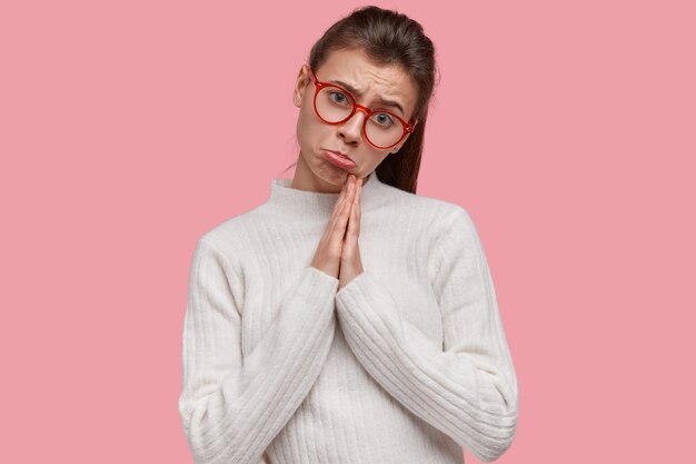 Retrato de mujer suplicante frunce los labios, mantiene las palmas de las manos en gesto de oración, pide fortuna, usa anteojos rojos, suéter blanco casual