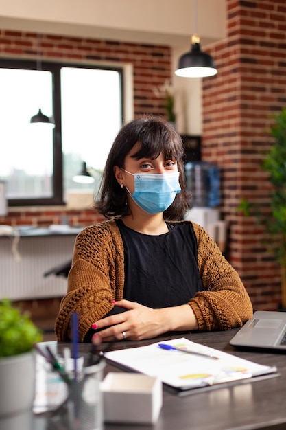 Retrato de mujer tenedora de libros con mascarilla de protección médica mirando a la cámara trabajando en la presentación de negocios revisando el informe financiero en la oficina de inicio. Manager analizando la estrategia de la empresa