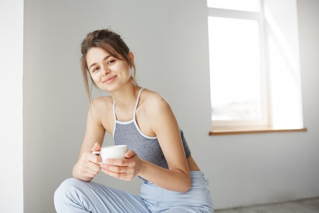 Retrato de mujer tierna joven sonriendo sosteniendo la taza sentado en la silla sobre la pared blanca temprano en la mañana.