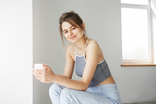 Retrato de mujer tierna joven sonriendo sosteniendo la taza sentado en la silla sobre la pared blanca temprano en la mañana.