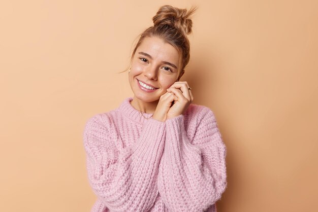 El retrato de una mujer tierna y linda mantiene las manos cerca de la cara, inclina la cabeza, tiene buen humor, sonríe con dientes, tiene el cabello peinado en un moño, usa un suéter de punto suelto aislado sobre un fondo beige, se ve con admiración