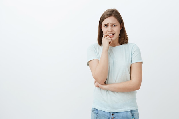 Retrato de mujer tímida ansiosa en traje casual frunciendo el ceño lloriqueando y mordiéndose la uña de los sentimientos nerviosos de pie inseguro y preocupado sobre la pared gris