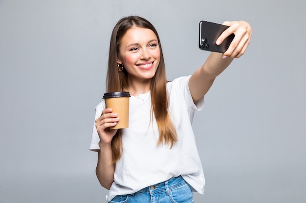 Retrato de mujer tomando fotos selfie en smartphone en la oficina y bebiendo café para llevar de un vaso de plástico aislado