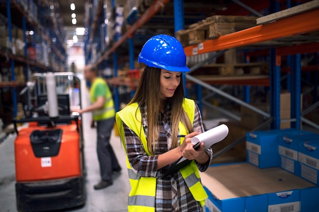 Retrato de mujer trabajadora de almacén comprobando el inventario en el departamento de almacenamiento mientras su compañero de trabajo operando la carretilla elevadora en segundo plano.