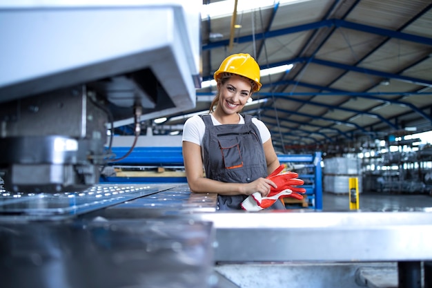 Retrato de mujer trabajadora de fábrica en uniforme protector y casco permanente por máquina industrial en la línea de producción