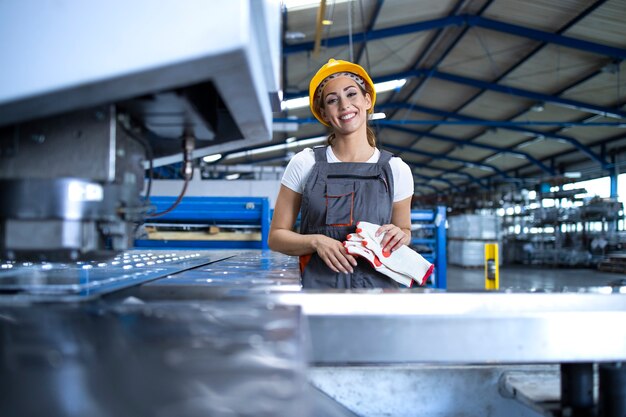 Retrato de mujer trabajadora de fábrica en uniforme protector y casco permanente por máquina industrial en la línea de producción