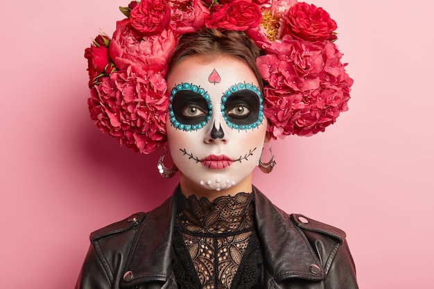 Retrato de mujer tranquila que celebra el Día de la Muerte, tiene maquillaje de calavera de azúcar, ojeras cerca de los ojos, sonrisa pintada, piensa que la muerte es parte natural del ciclo humano, viste atuendo tradicional mexicano.