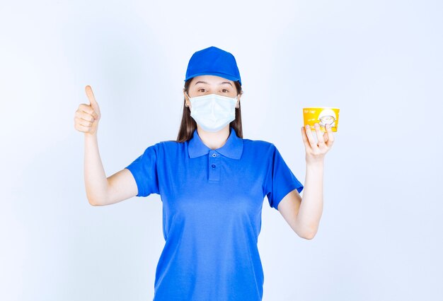 Retrato de mujer en uniforme y máscara médica con vaso de plástico mostrando el pulgar hacia arriba