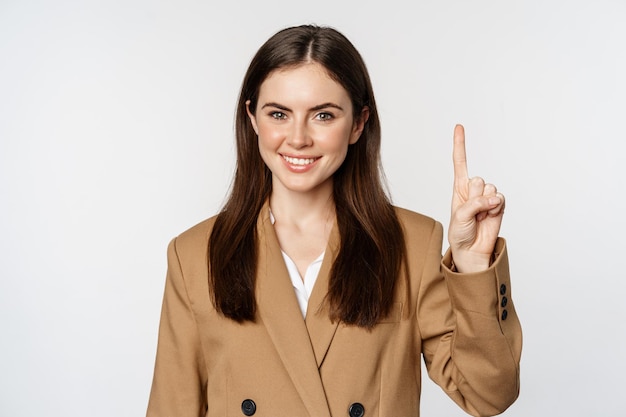 Retrato de mujer vendedora corporativa mostrando el dedo número uno y sonriendo de pie en traje sobre w...