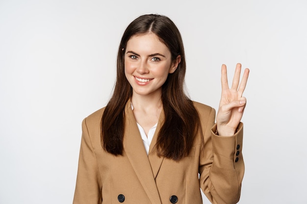 Retrato de mujer vendedora corporativa mostrando el número tres dedos y sonriendo de pie en traje sobre...