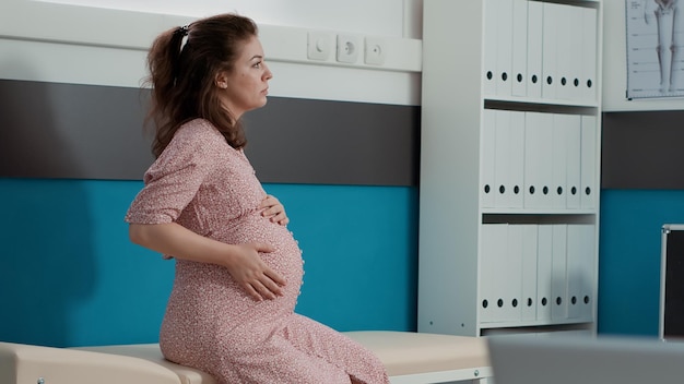 Retrato de mujer con vientre de embarazo esperando para iniciar cita de consulta con especialista en salud. Futura madre esperando un hijo y asistiendo a un examen de chequeo en el gabinete médico.