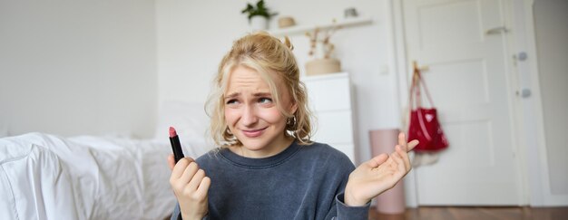 Retrato de una mujer vlogger que parece decepcionada mostrando lápiz labial y encogiéndose de hombros grabando