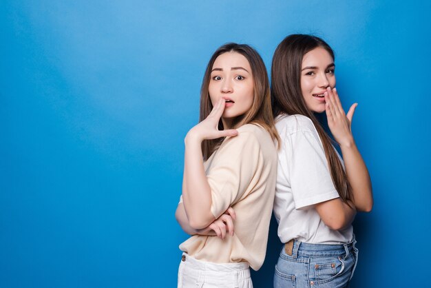 Retrato de mujeres emocionadas multinacionales en ropa casual sonriendo y tapándose la boca aislada sobre pared azul