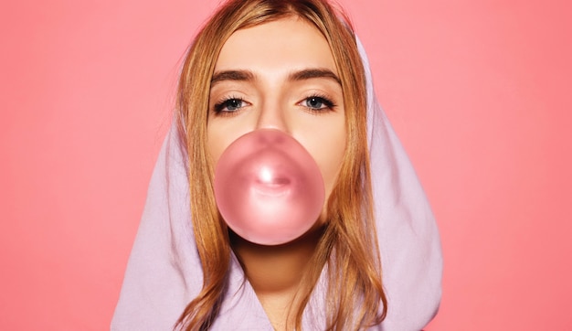 El retrato de mujeres rubias juguetonas jovenes se vistió en la sudadera con capucha azul que se colocaba aislada sobre la pared rosada mientras que soplaba la burbuja con el chicle. mujer