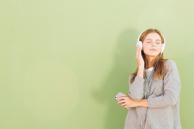 Retrato de una música que escucha de la mujer bastante joven en el auricular a través del teléfono móvil contra el contexto del verde menta