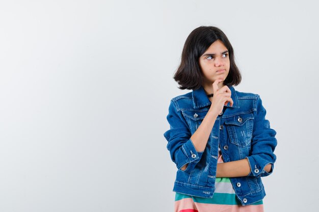 Retrato de niña adolescente de pelo medio cortado en camiseta colorida y abrigo de mezclilla puso la mano en la barbilla y el pensamiento aislado sobre fondo blanco.