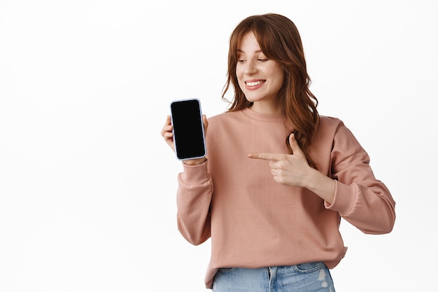 Retrato de niña adolescente sonriendo, señalando con el dedo y mirando la pantalla del teléfono, mostrando la aplicación en el teléfono móvil, la pantalla del teléfono inteligente, de pie en blanco