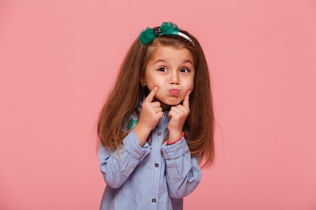 Retrato de niña adorable con hermoso cabello castaño largo explotando sus mejillas tocando la cara