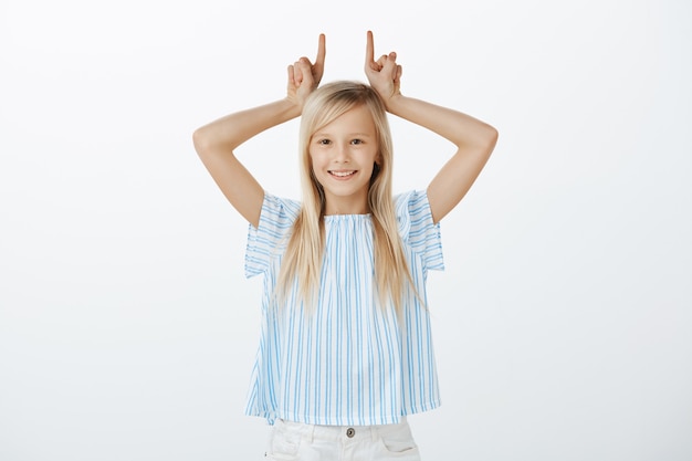 Retrato de niña adorable juguetona con cabello rubio, divirtiéndose mientras se burla, sosteniendo los dedos índices sobre la cabeza como si mostrara cuernos, sonriendo con alegría sobre la pared gris