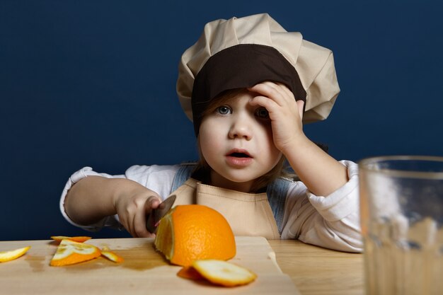 Retrato de niña adorable en sombreros de chef y delantal cortando naranjas en la placa de cocina con un cuchillo, haciendo jugo de cítricos frescos o un desayuno saludable. Concepto de vitamina, frescura, dieta y nutrición.