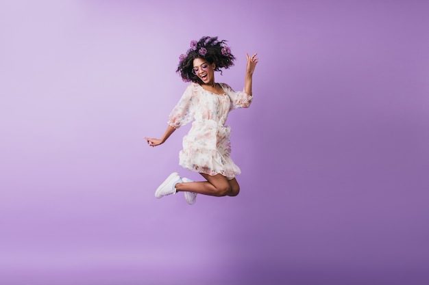 Retrato de niña africana divertida en traje blanco saltando. alegre joven morena que expresa emociones positivas.