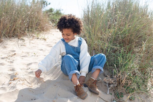 Retrato de niña afroamericana en la playa. Modelo femenino con cabello rizado en jeans en general sentado en la arena, jugando. Retrato, concepto de belleza