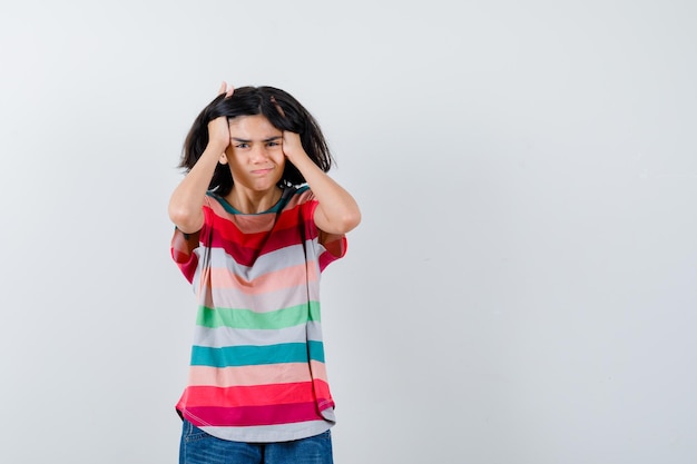 Retrato de niña agarrando la cabeza con las manos en camiseta y mirando irritada vista frontal