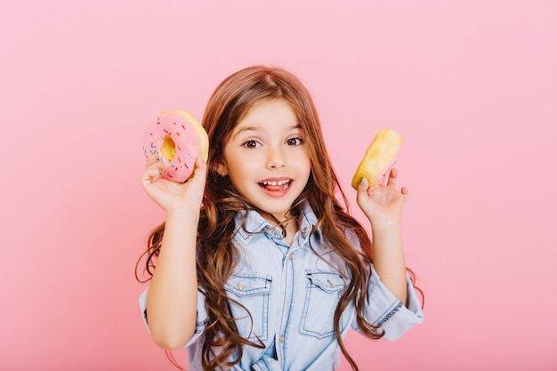 Retrato de niña alegre con cabello largo morena divirtiéndose a la cámara con coloridos donuts aislado sobre fondo rosa. Expresando verdaderas emociones positivas de niño lindo. Lugar para el texto