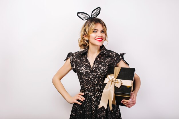 Retrato de niña alegre con cabello largo y rizado morena, vestido negro de lujo. Celebrando, fiesta de cumpleaños, divirtiéndose con caja de regalo.