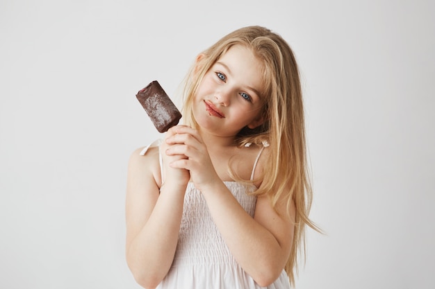 Retrato de niña alegre con ojos azules y cabello claro disfrutando de su helado con restos de chocolate en su rostro. Infancia feliz y despreocupada