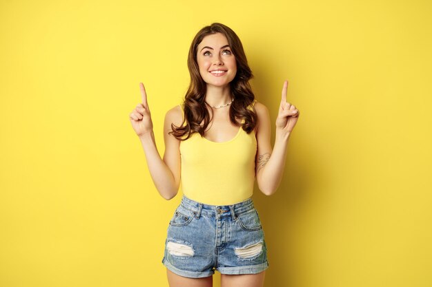 Retrato de niña alegre sonriendo, señalando con el dedo hacia arriba, mostrando texto promocional, anuncio de verano, de pie contra el fondo amarillo