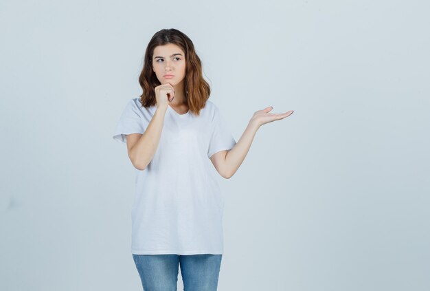 Retrato de niña apoyando la barbilla en la mano, extendiendo la palma hacia un lado en una camiseta blanca y mirando pensativa vista frontal