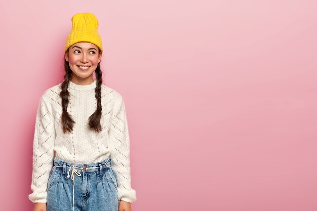 El retrato de una niña asiática feliz se ve con alegría a un lado, tiene una sonrisa agradable, vestida con un sombrero amarillo, suéter blanco y jeans, tiene mejillas rojas y maquillaje, posa sobre una pared rosada