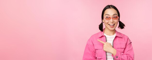 Retrato de niña asiática sonriente en traje elegante gafas de sol apuntando con el dedo a la izquierda mostrando publicidad