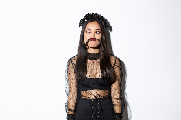 Retrato de niña asiática tonta y divertida tonteando, haciendo muecas y haciendo bigote de hebra de cabello, de pie sobre fondo blanco en disfraz de halloween.