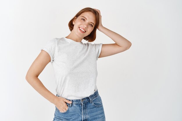 Retrato de niña de belleza natural con maquillaje ligero, tocando el pelo corto y sonriendo feliz, de pie en camiseta casual con jeans contra la pared blanca