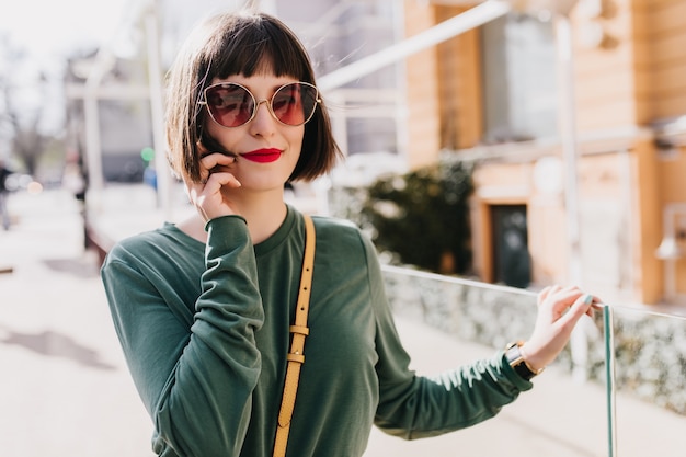 Retrato de niña blanca interesada con pelo corto posando con gafas de sol oscuras. Foto al aire libre de mujer elegante dichosa en suéter verde escalofriante en fin de semana.