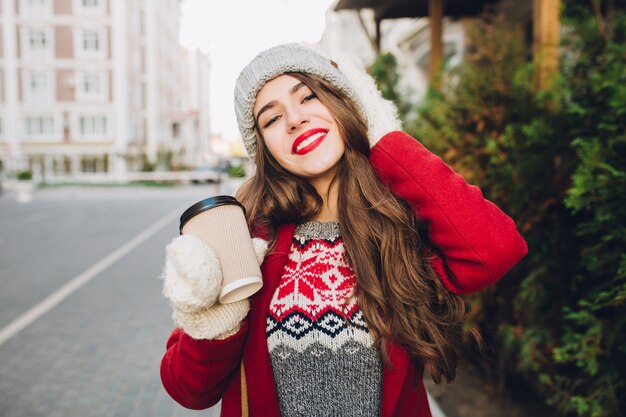 Retrato de niña bonita en abrigo rojo y gorro de punto caminando en la calle. Ella sostiene el café para llevar con guantes blancos, sonriendo amigablemente con labios rojos.