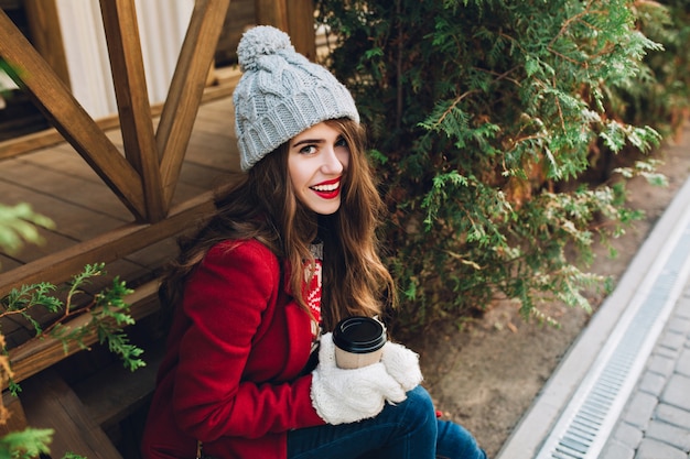 Retrato de niña bonita en abrigo rojo, gorro de punto sentado en escaleras de madera cerca de ramas verdes al aire libre. Ella sostiene café con guantes blancos y sonriendo. Vista desde arriba.