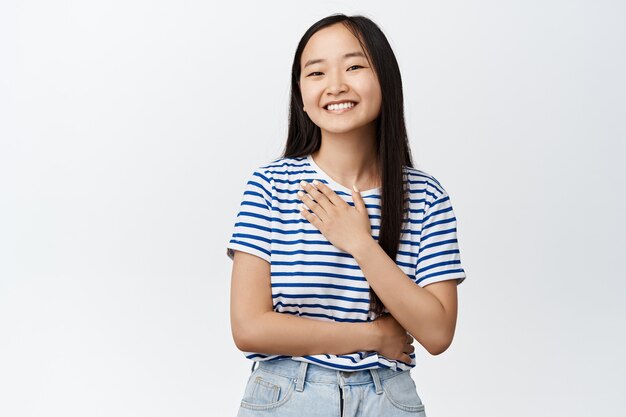 Retrato de niña bonita asiática con rostro hermoso natural, sonriendo feliz, sosteniendo la mano en el pecho halagado, expresando aprecio, de pie en blanco