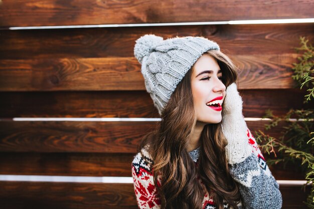 Retrato de niña bonita con cabello largo y labios rojos en gorro de punto y guantes calientes en madera. Ella está sonriendo a un lado.