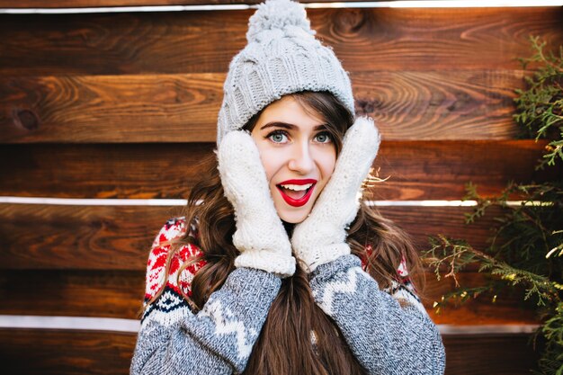 Retrato de niña bonita con cabello largo y labios rojos en gorro de punto en madera. Ella está tocando la cara con guantes calientes, sonriendo.