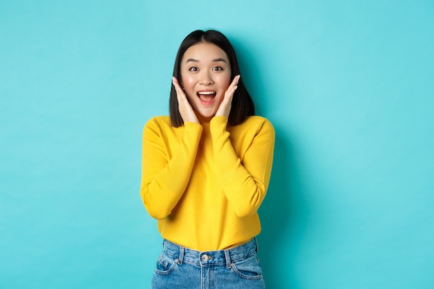 Retrato de niña bonita coreana recibe noticias sorprendentes, mirando sorprendido y feliz a la cámara, de pie sobre fondo azul.