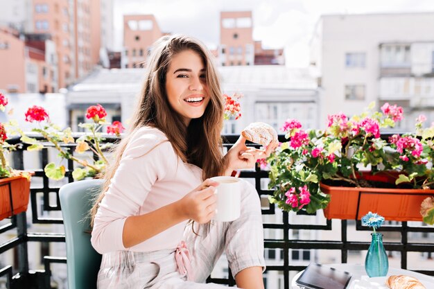 Retrato de niña bonita desayunando en el balcón rodean flores en la soleada mañana en la ciudad. Ella sostiene una taza, croissant, sonriendo.