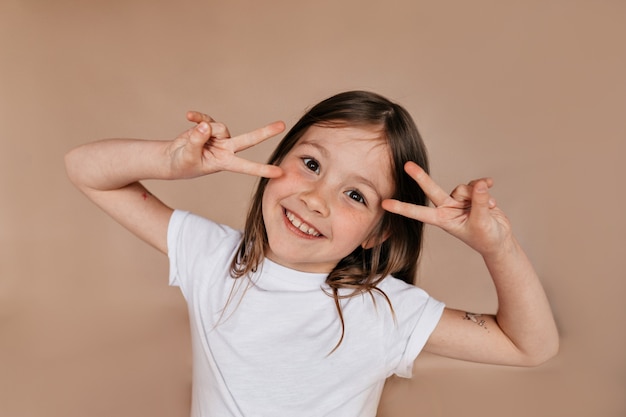 Retrato de niña bonita encantadora mostrando signos de paz cerca de la cara y sonriendo sobre pared beige